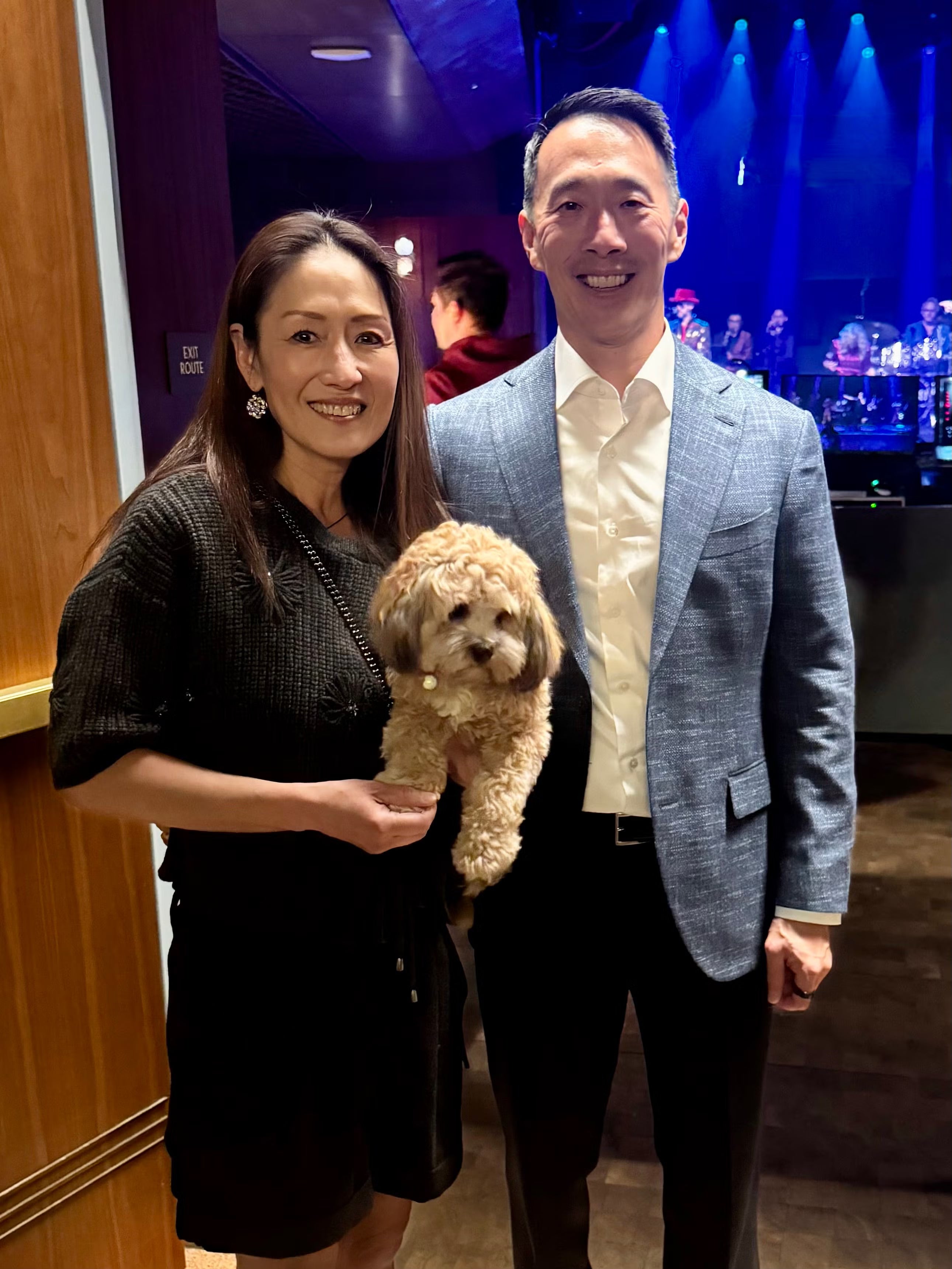 Smiling couple at dim venue; woman holds small curly tan dog with blue stage lights behind