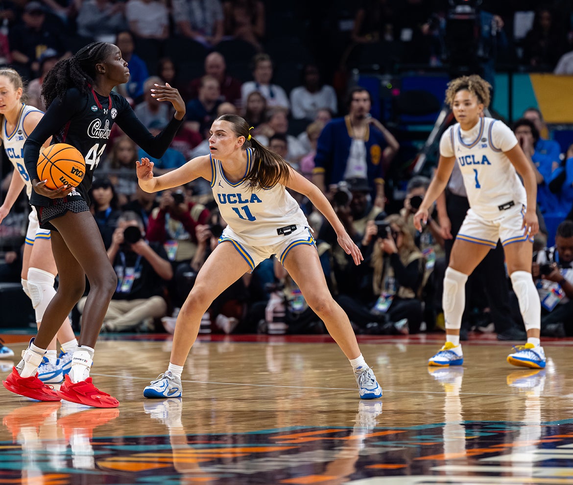 Two UCLA basketball players watch and guard as a South Carolina player dribbles down the court