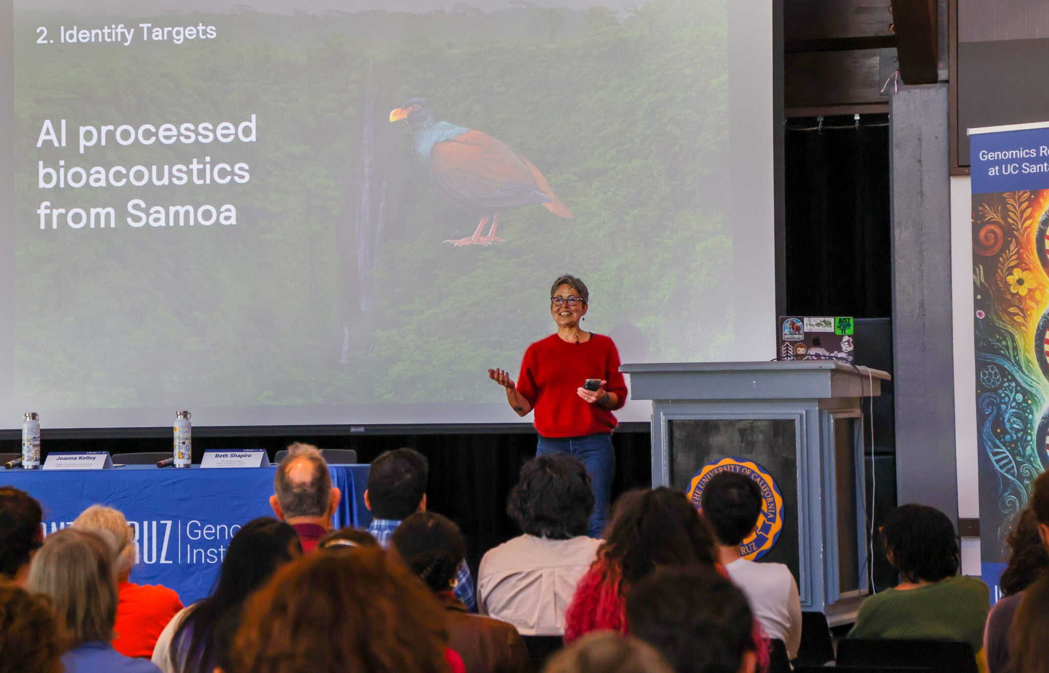 A woman in a red sweater speaks to a crowd in front of a large projector screen with a toucan projected on it