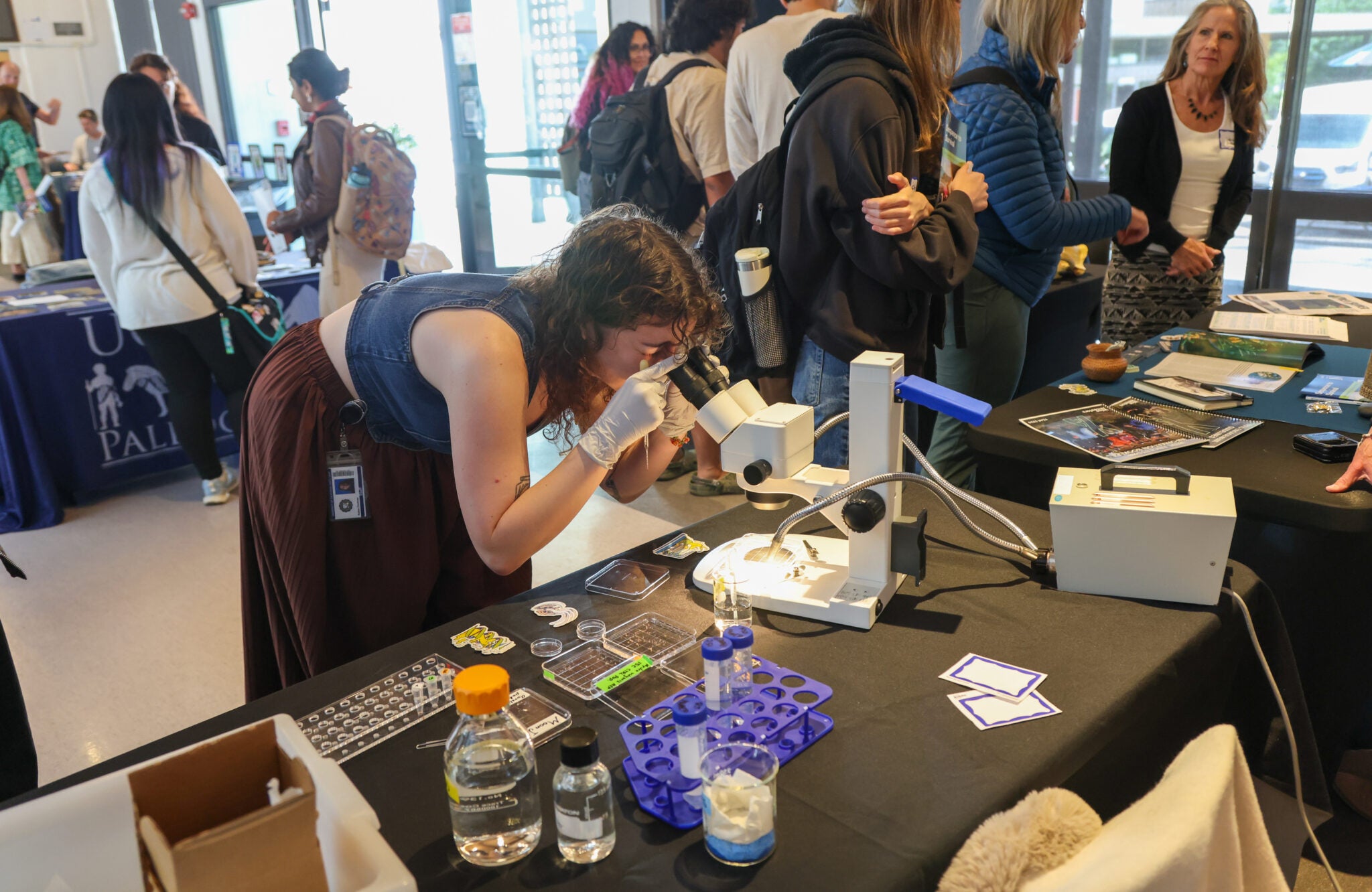 A woman with gloves on peers into a microscope that is set up at a booth. Other symposium participants talk around her.