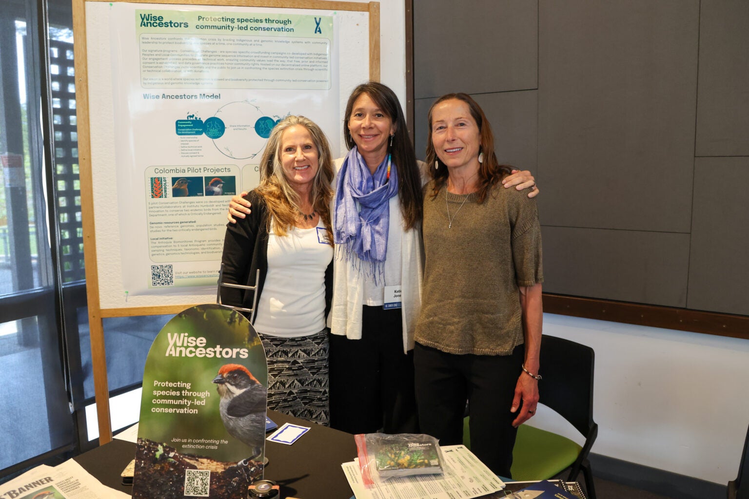 Three women pose behind a sign that reads Wise Ancestor