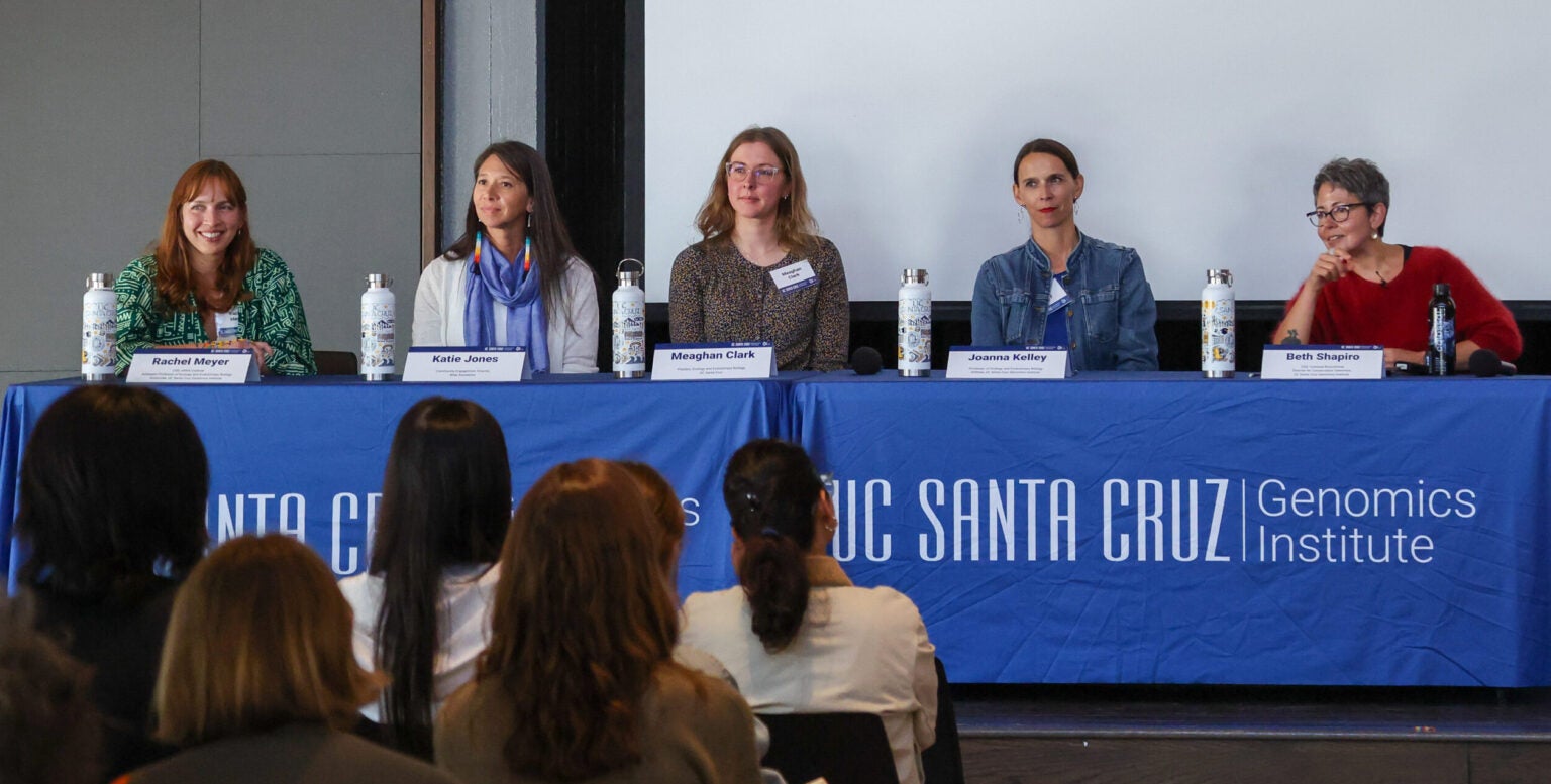 Five women sit facing the audience behind a table with a bright blue tablecloth labeled UC Santa Cruz Genomics Institute