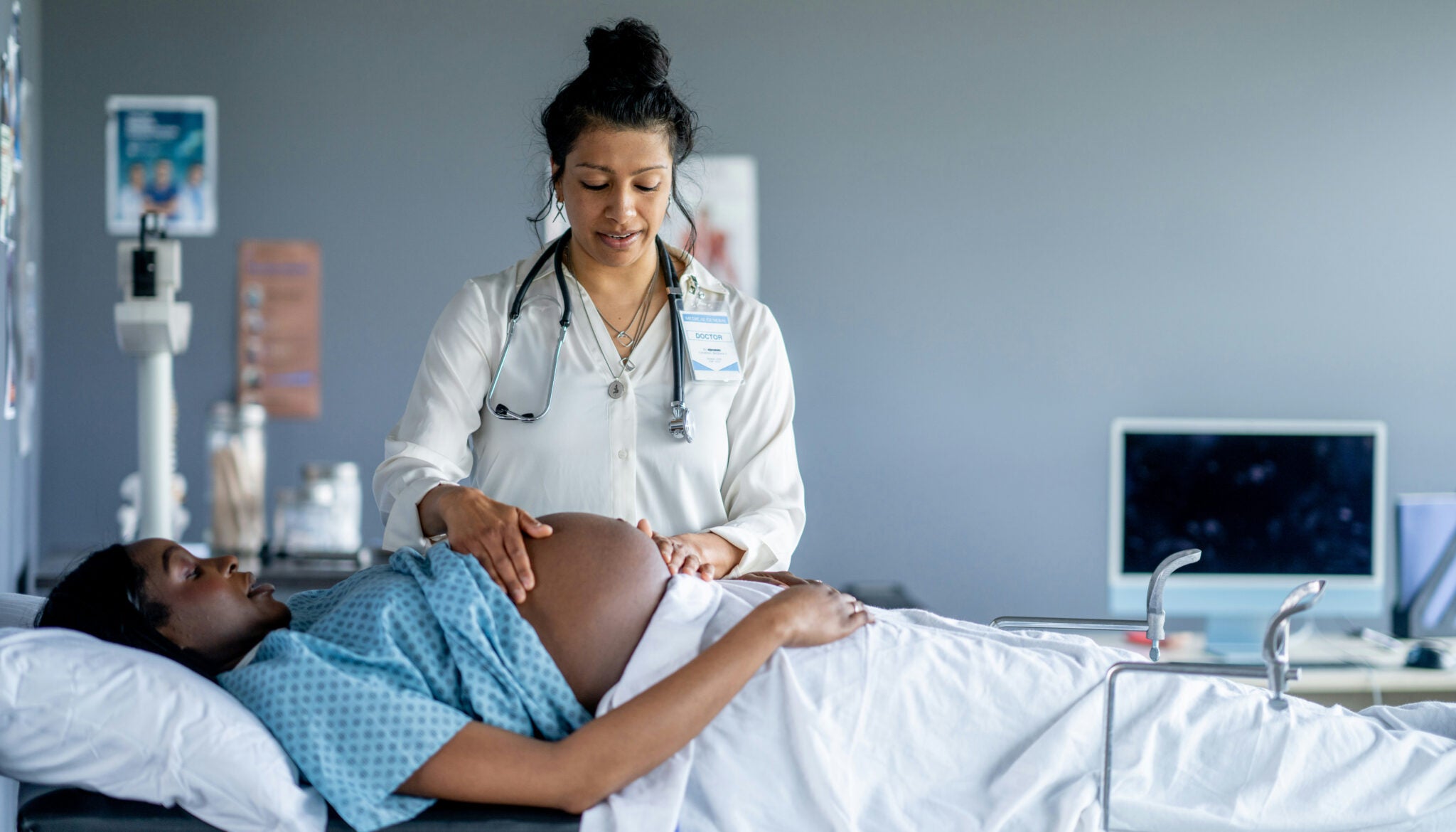 A female doctor in a medical setting palpitates the exposed belly of a pregnant woman who's lying on a bed