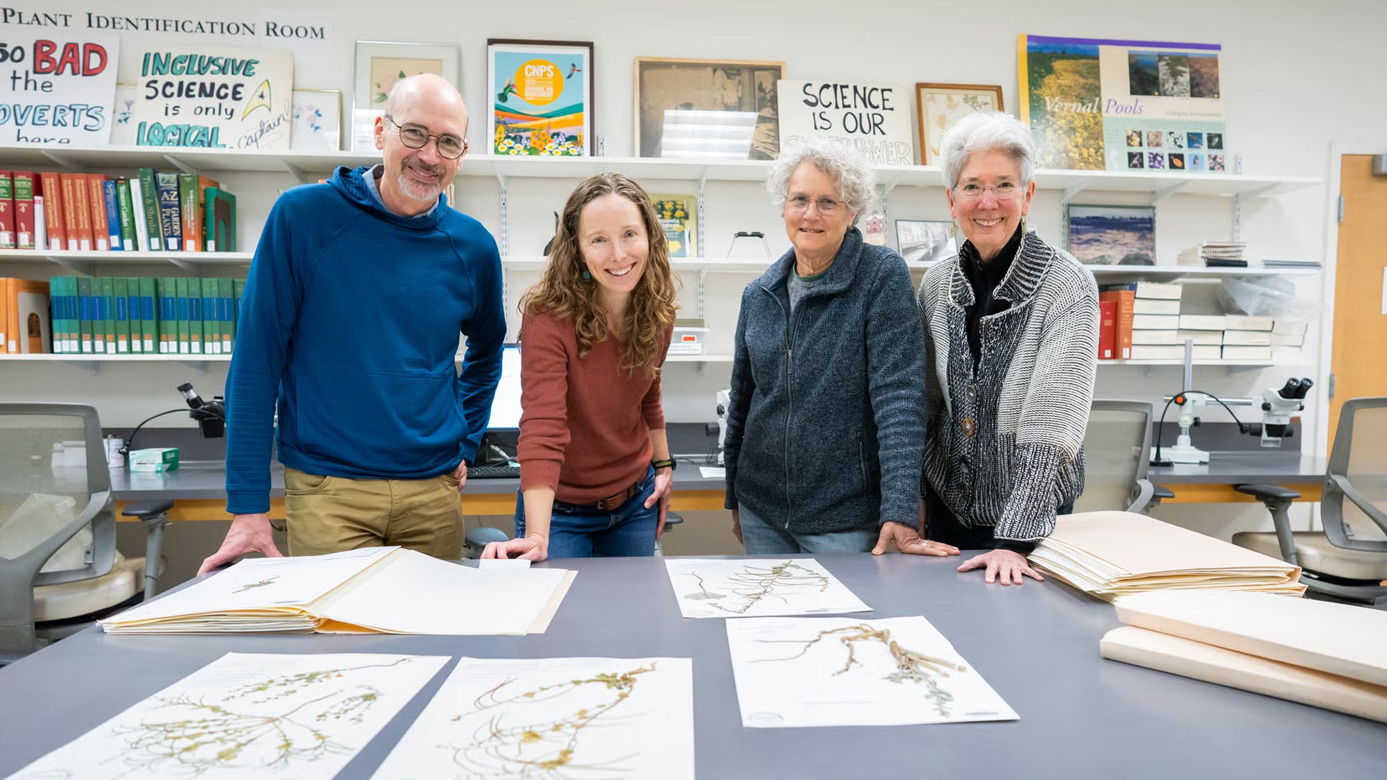 Four people smiling, leaning on a table with botanical prints in the forground