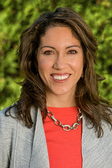 A woman smiles in a headshot while wearing a gray blazer and orange-red top