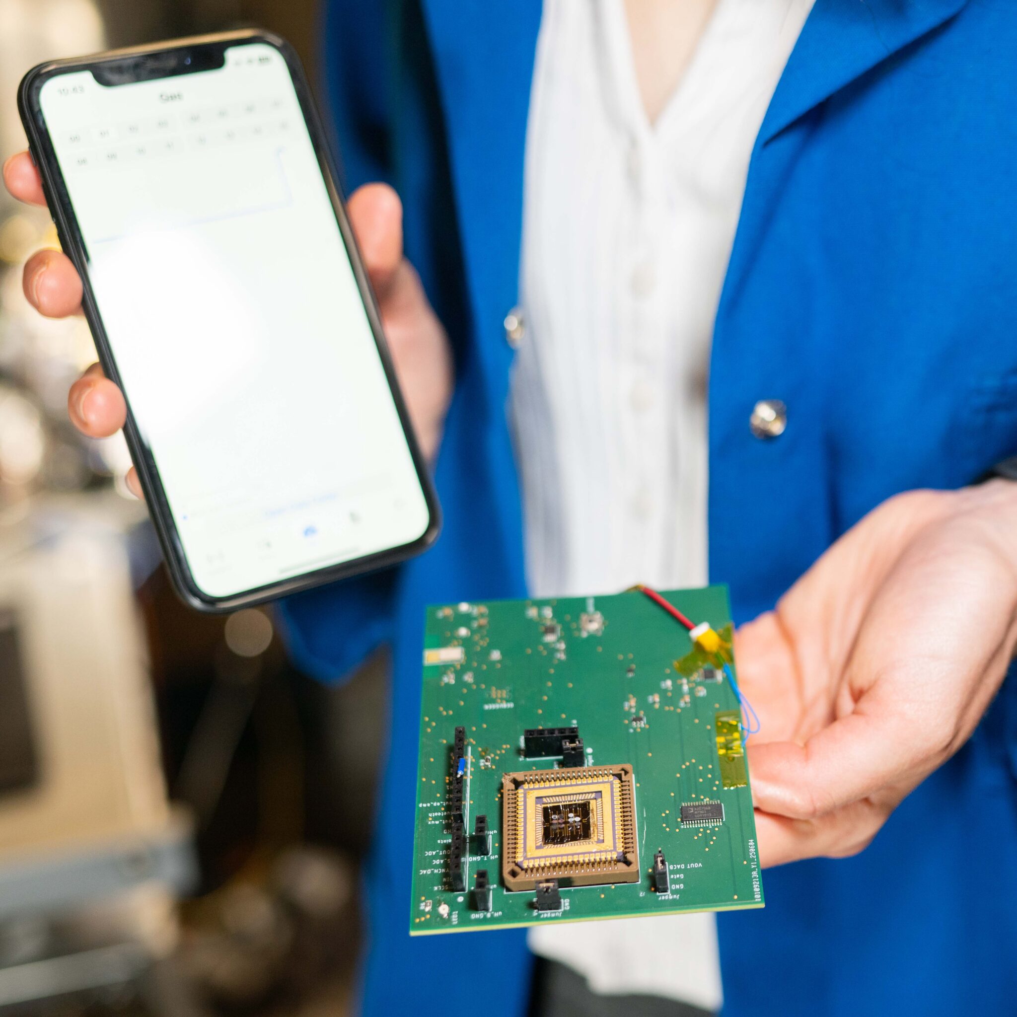 A person holds an iPhone in one hand and a green electronic board with the electronic nose attached in the other hand