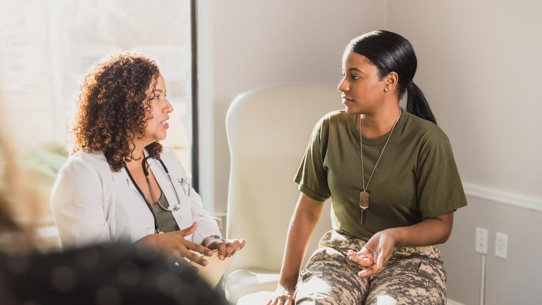 A woman in a medical coat speaks to a soldier in a green shirt and camo pants