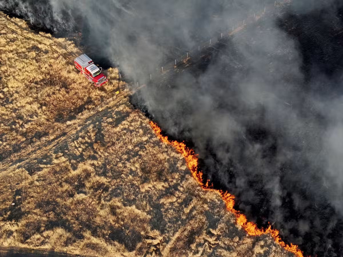 A fire truck works at the edge of a spreading grass fire, one half of the field blackened with smoke and burns