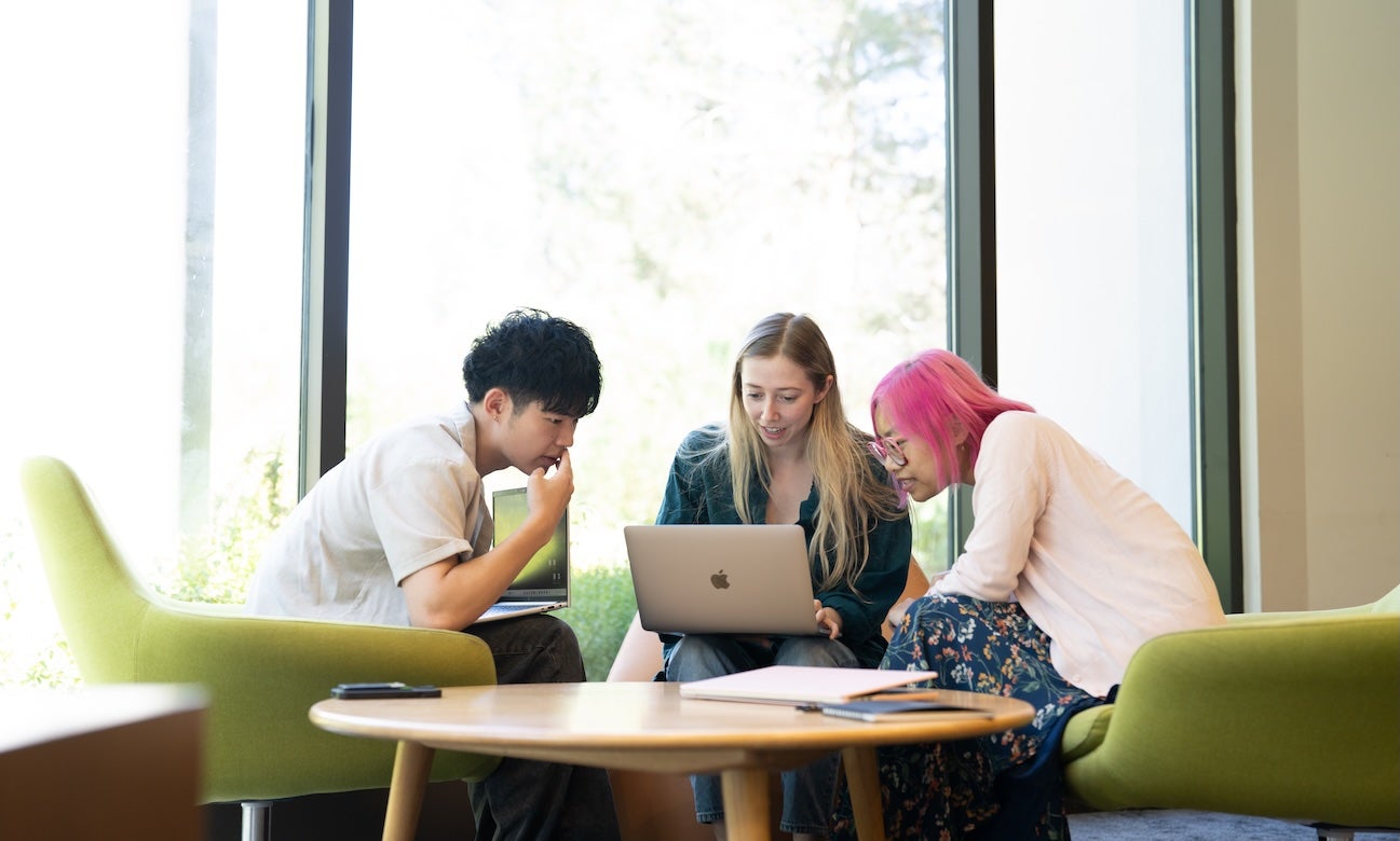 Three students in a lounge with green chairs lean in and look at a computer screen