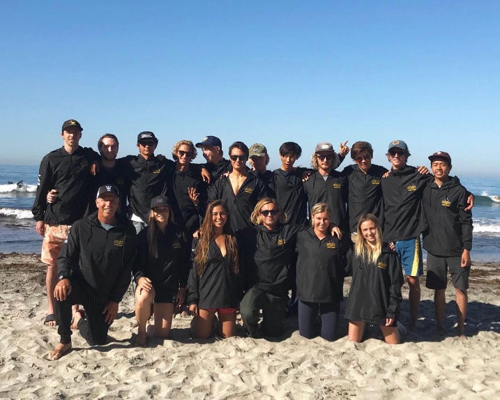 UC San Diego surf team poses for a photo in 2 rows, wearing matching black tops, on a beach on a sunny day