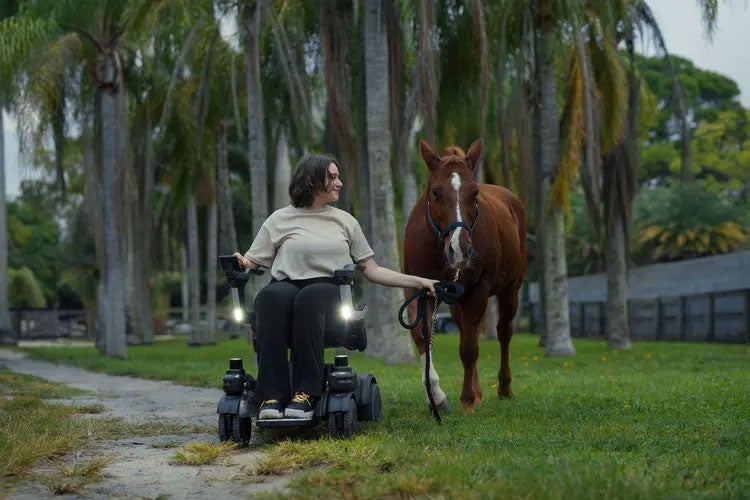 A woman seated in a powered wheelchair stands beside a horse on a paved path.