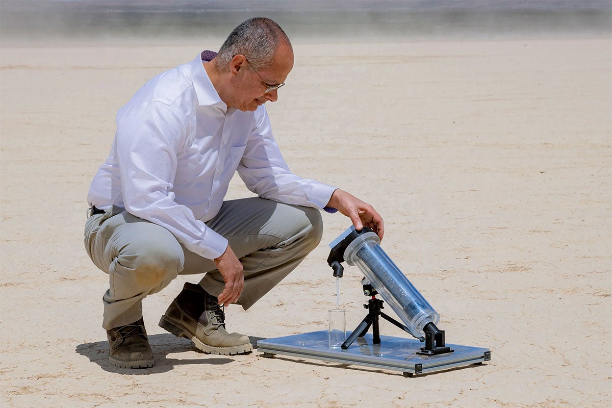 A man crouches beside a cylindrical device on a tripod in a sandy landscape, with a small glass beneath it.