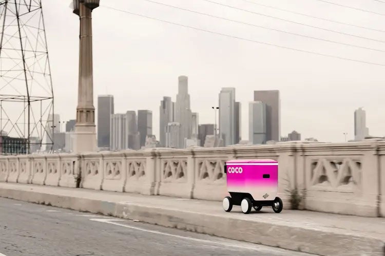 A small pink cooler on wheels rolls along a city sidewalk.