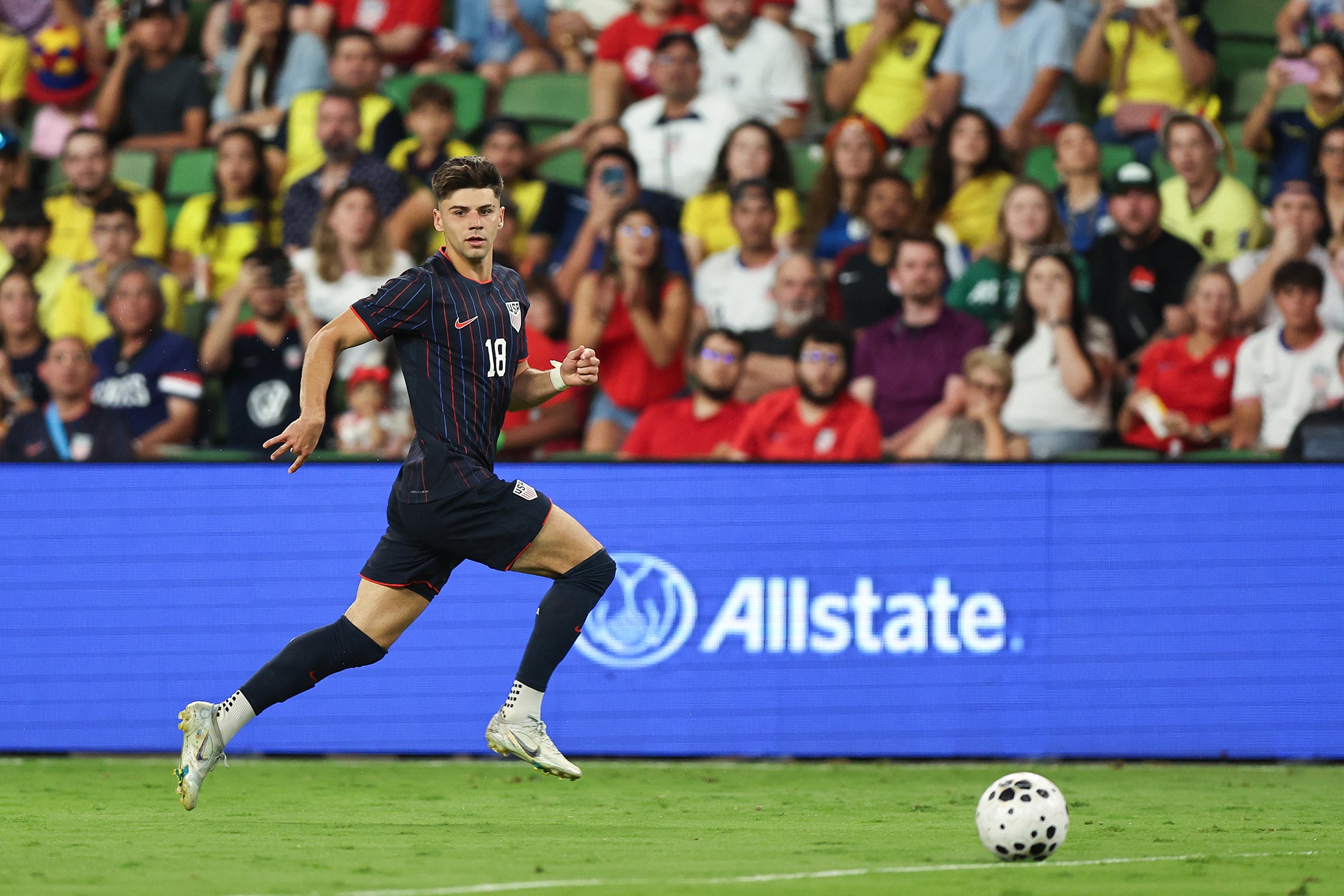Max Arfsten of the United States in a navy blue uniform with stripes drives a ball during an international friendly match in front of the crowd, looking toward the camera
