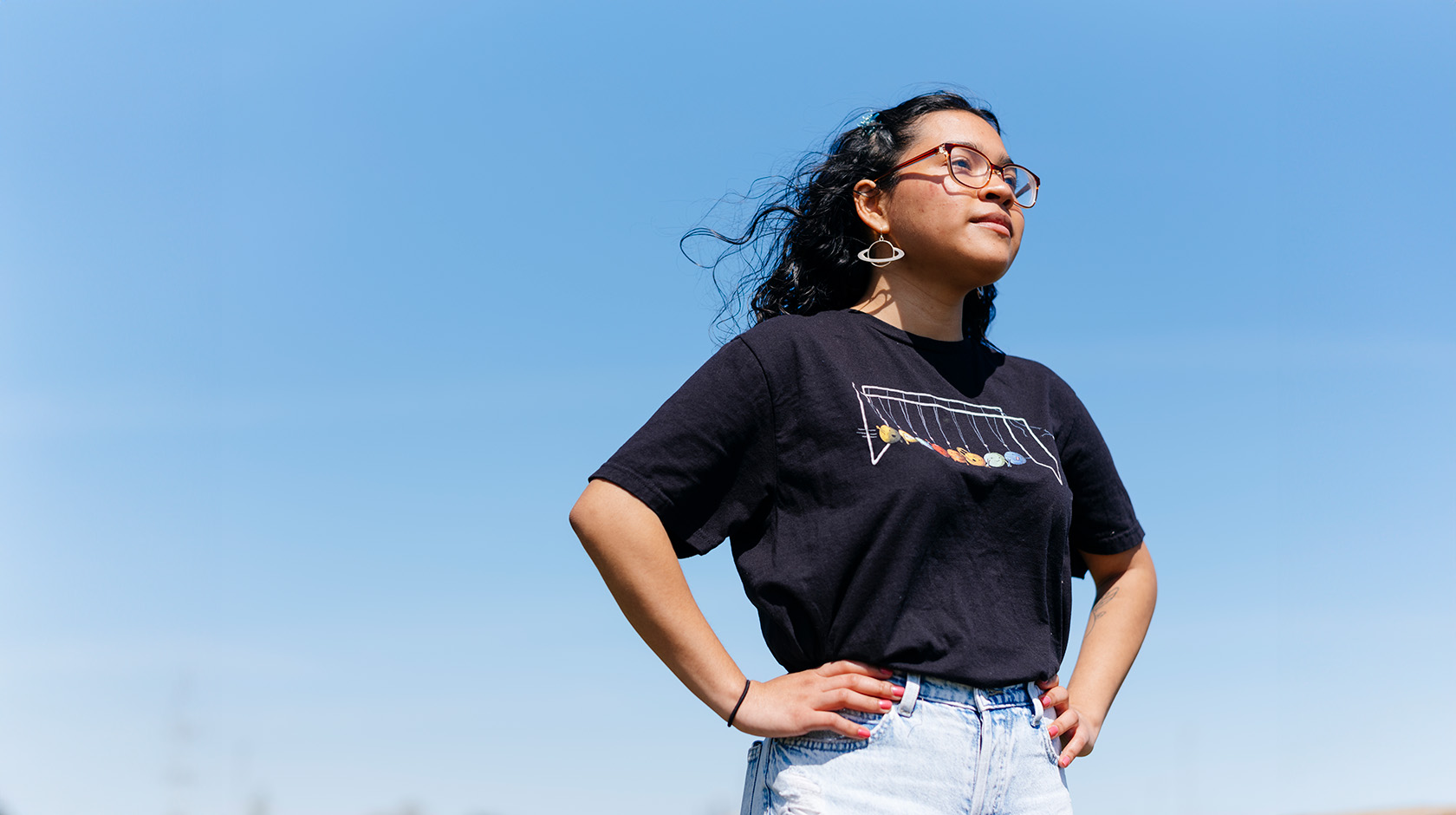 A young woman in a dark t-shirt, jeans and dangly earrings stands with her hands on her hips, looking determinedly forward, shot slightly from below, against a clear blue sky