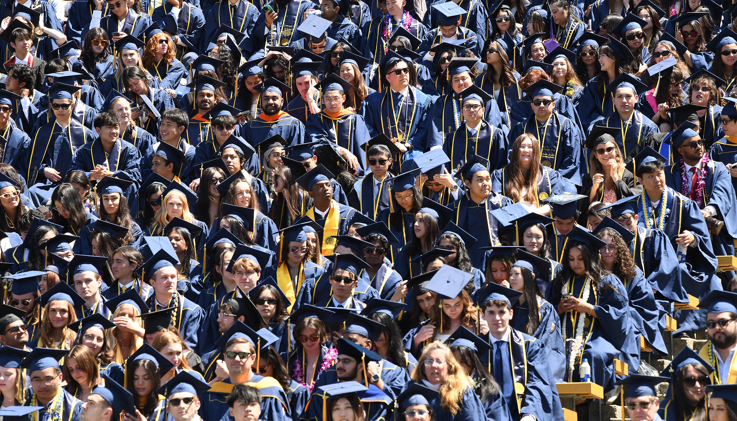 A high-angle shot of a dense crowd of university graduates in blue caps and gowns filling stadium bleachers, accented by colorful leis, stoles and sunglasses under bright sunlight.