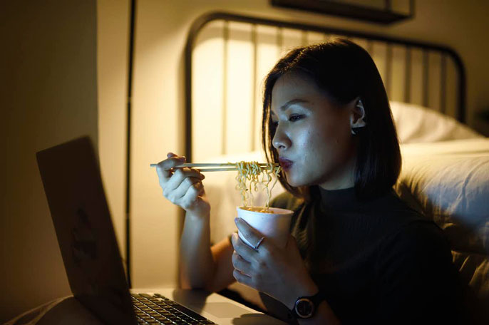 A young woman eating noodles at night watching something on the computer