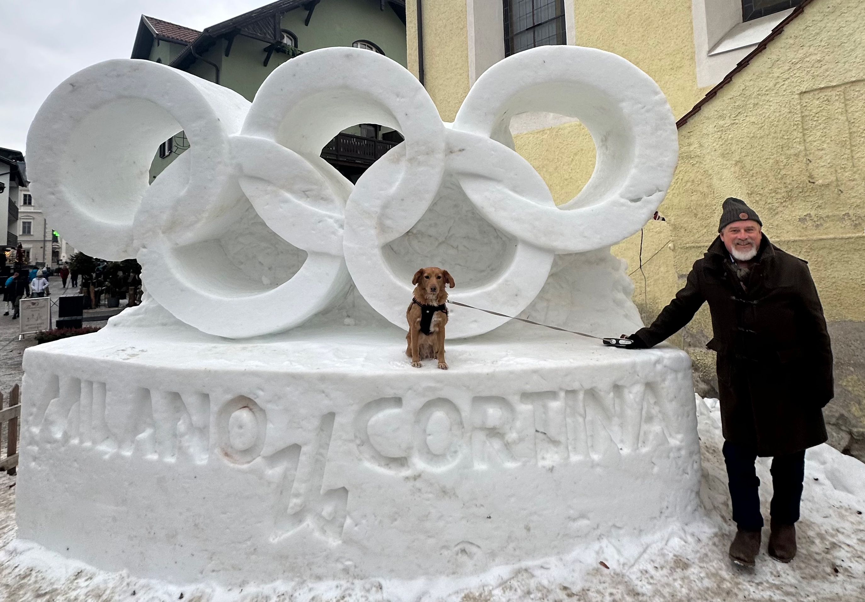In front of a snow sculpture of the Olympic rings that reads Milano Cortina, a man stands next to a service dog sitting on the sculpture