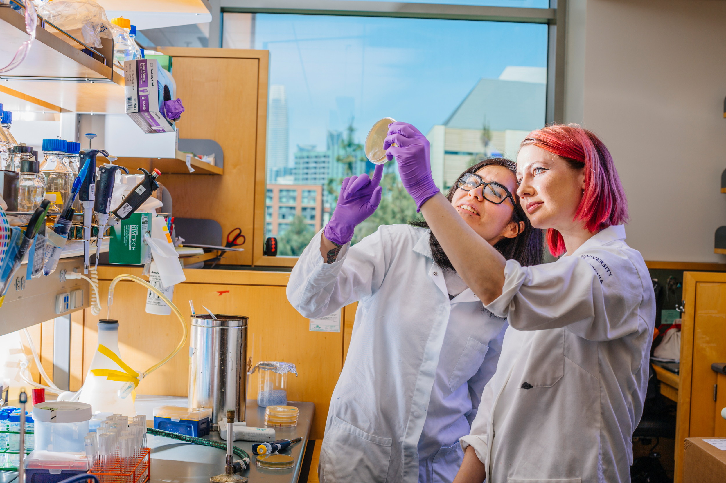 Two female students, one with pink hair, hold up a petri dish in a lab