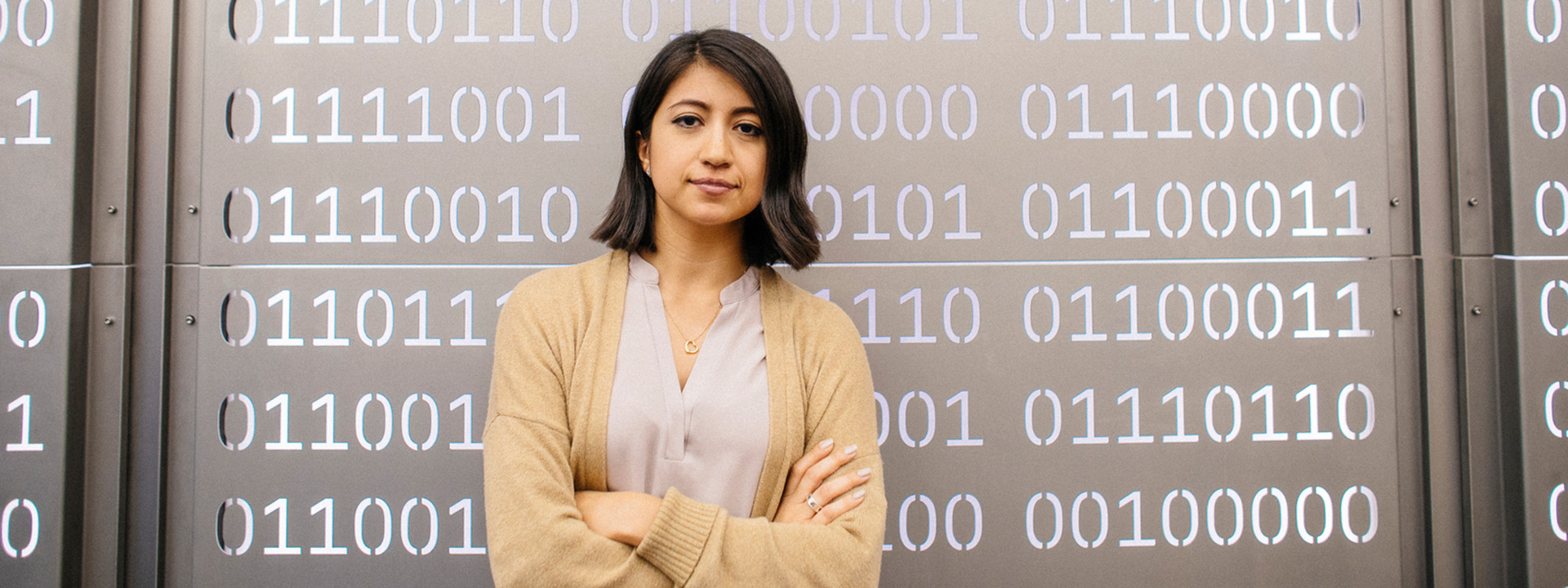 A woman with shoulder length dark hair wearing a cream colored cardigan looks resolutely at the camera with her arms crossed. She's standing in front of a metal panel with sequences of 0s and 1s cut out to simulate computer code. 