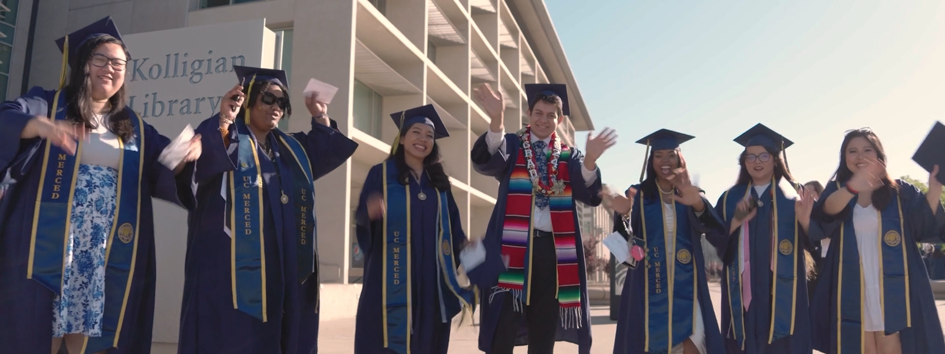 A row of students in graduate robes, caps and stoles wave hands excitedly in front of a library