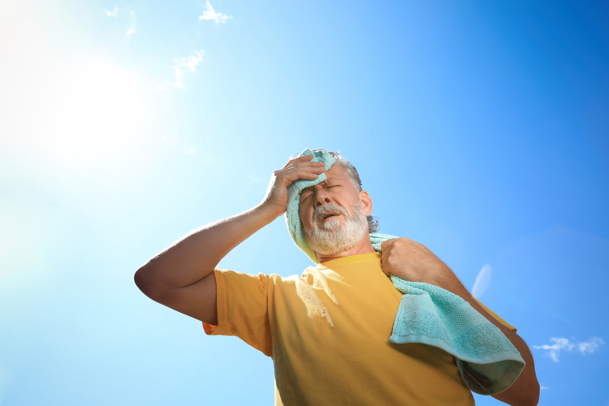 An older man wipes sweat off his brow, carrying a towel outside, bright sun overhead