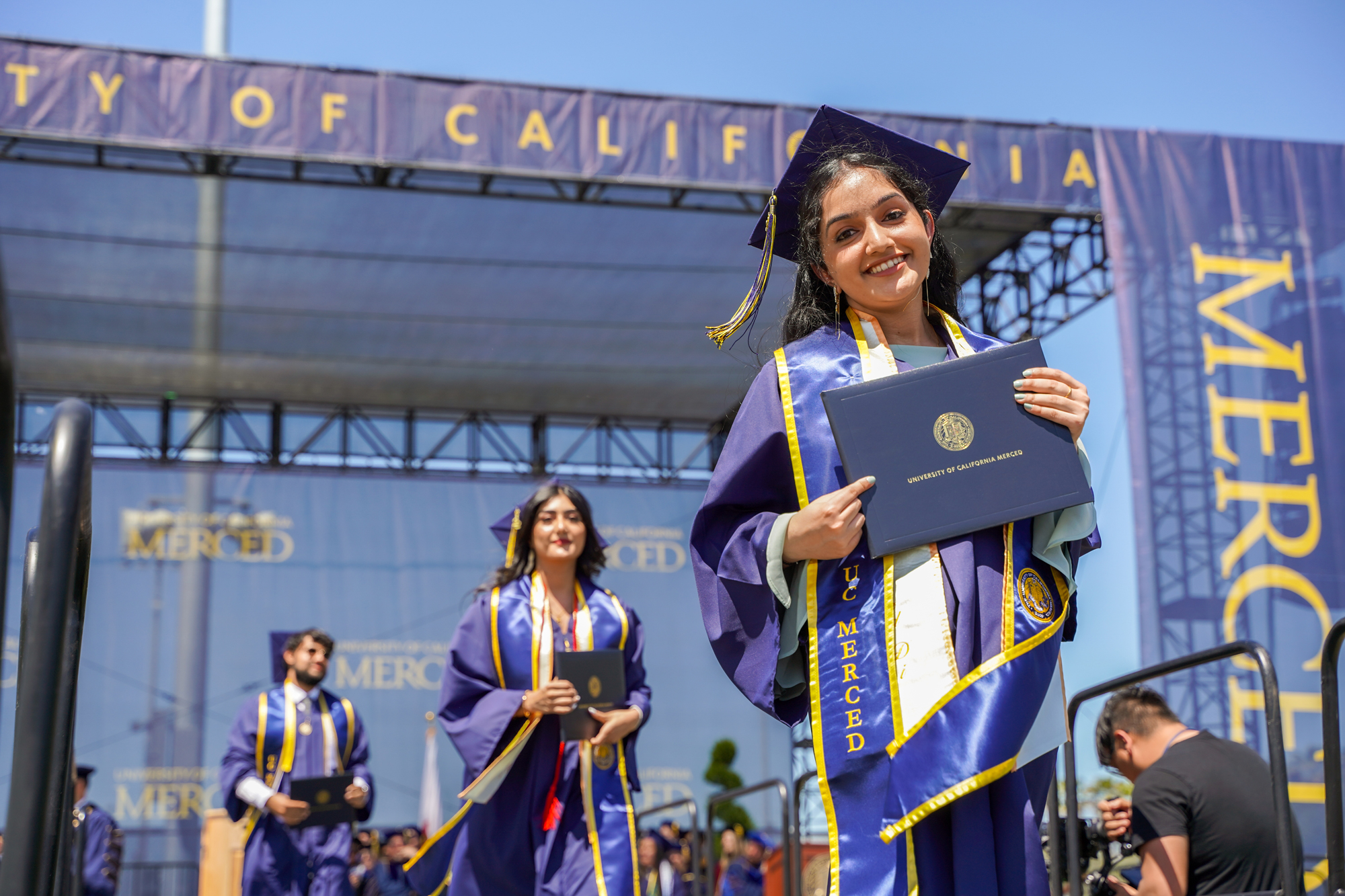 Three UC Merced graduates hold their diplomas as they leave the stage of graduation, smiling