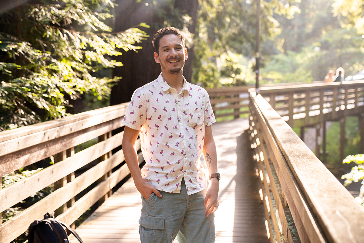 A smiling young man standing on a bridge surrounded by redwood trees