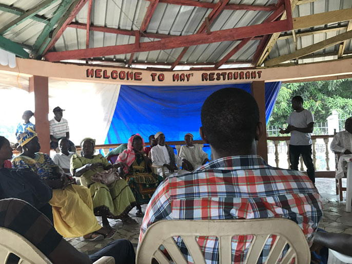 People sit in a circle and listen to a man talk on stage at an outdoor restaurant