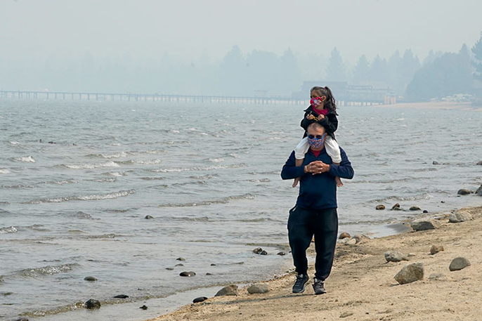 A man and his child walk along a shoreline in masks during smoky weather