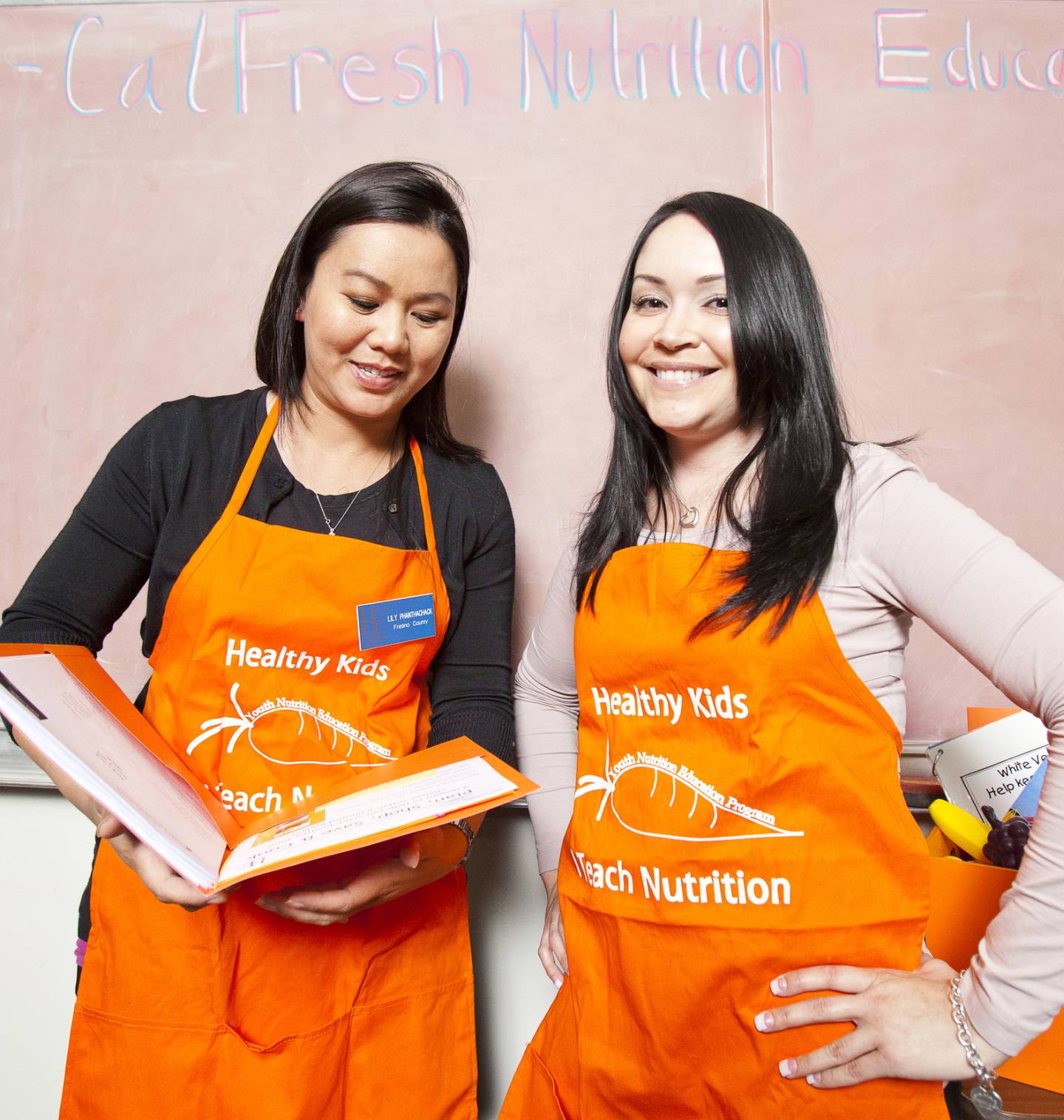 Two women in orange aprons one holding a book