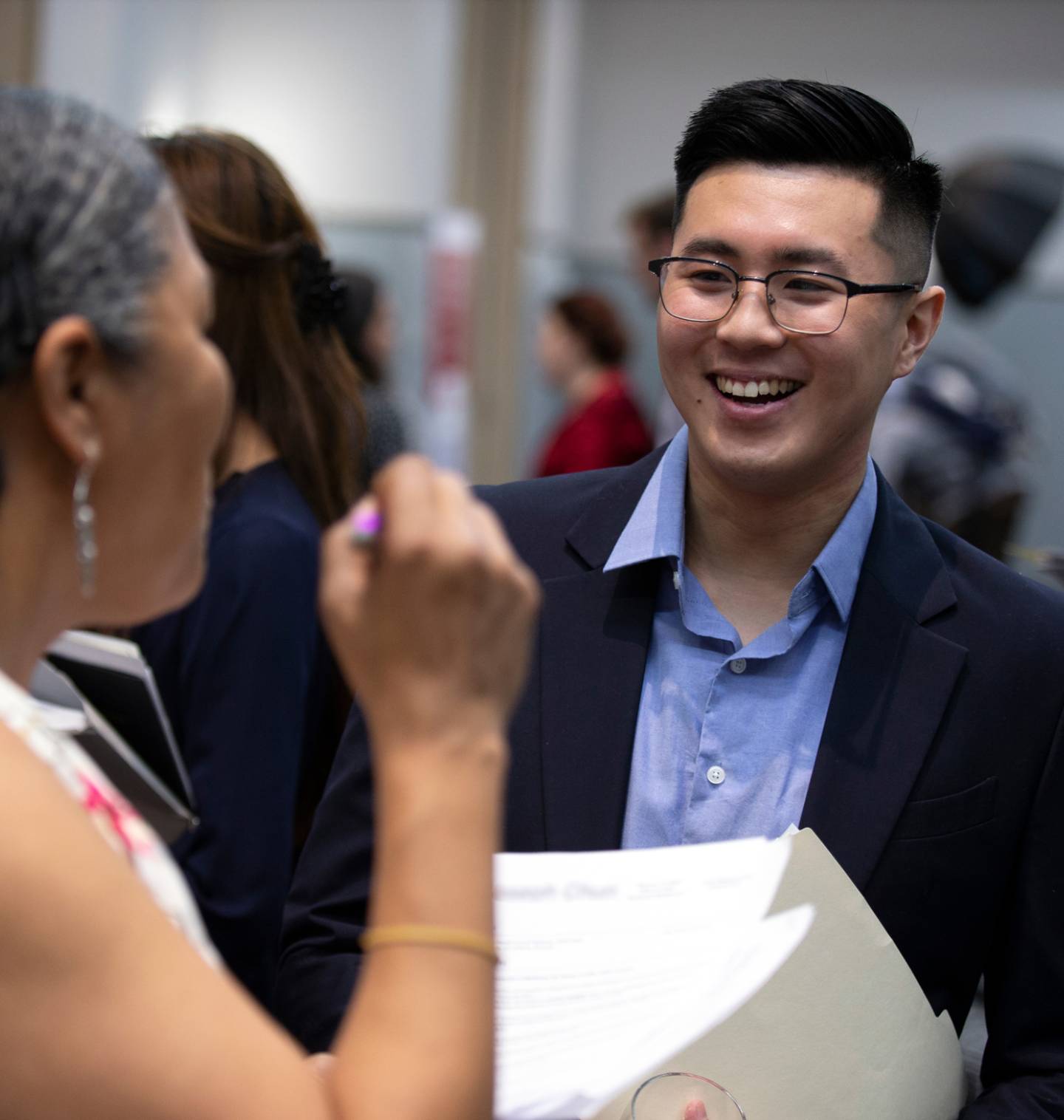 A middle-aged Black woman talks excitedly to a young Asian man in professional clothes holding his resume