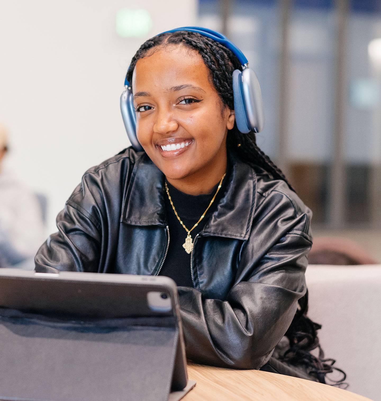 A young woman wearing blue over-ear headphones and a black leather jacket, smiles at the camera while sitting at a table with a tablet.