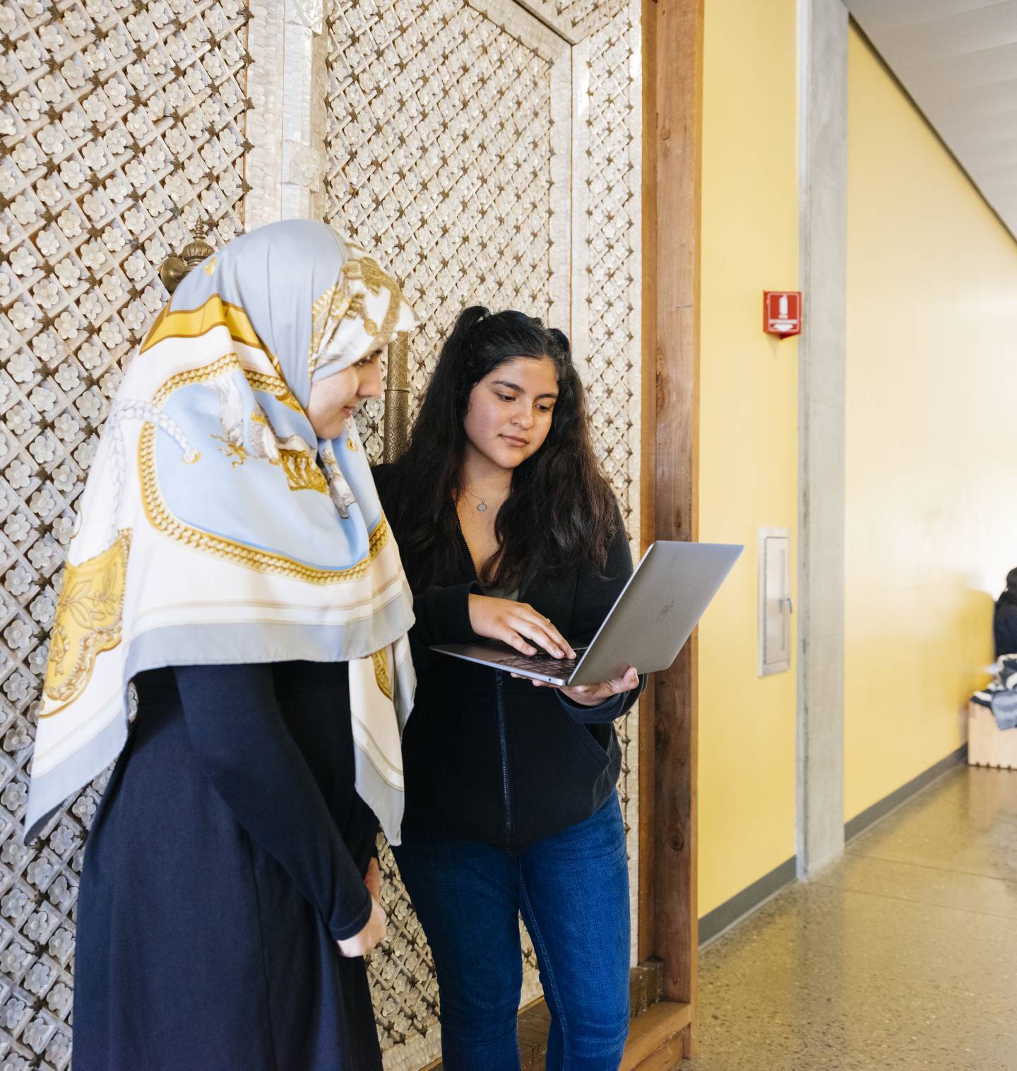 Two women one in a head scarf look at a computer together in a hallway