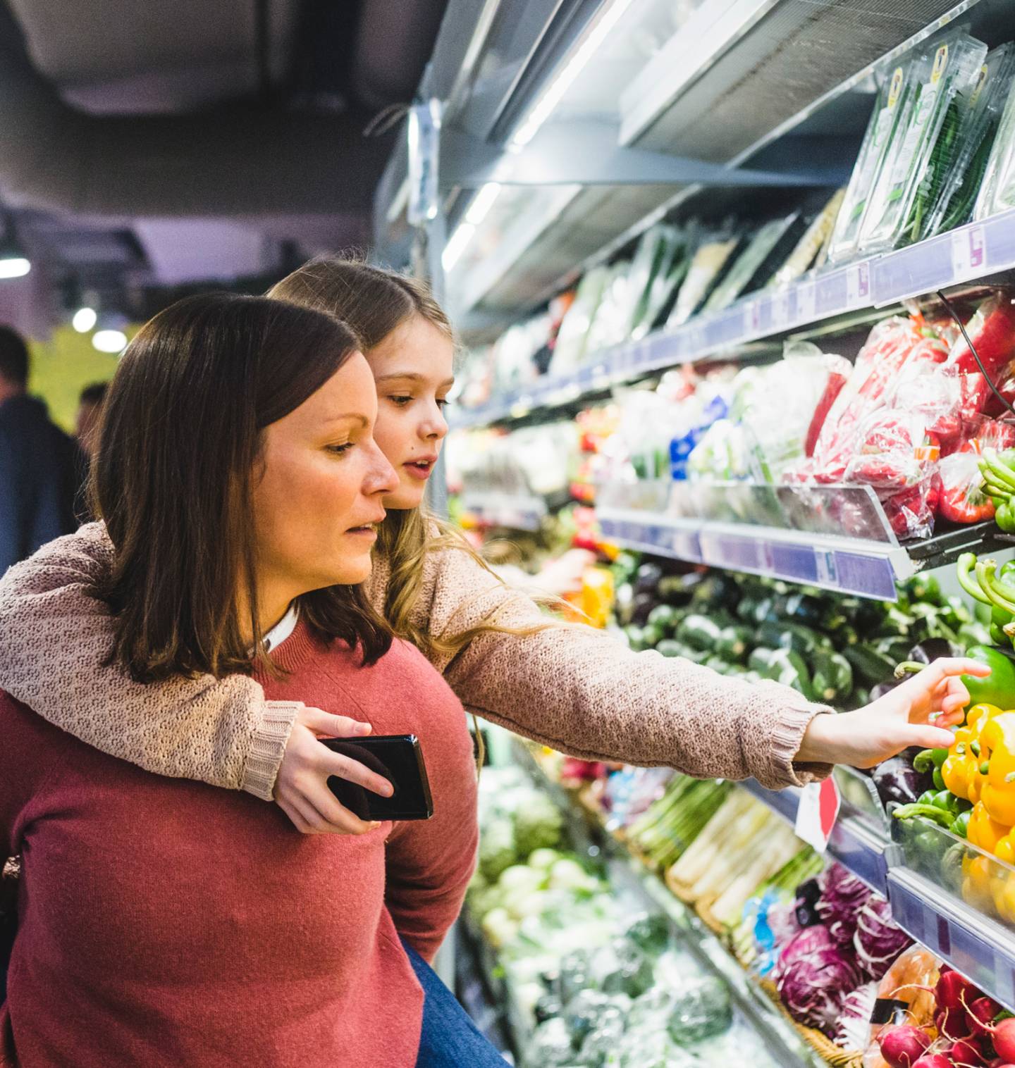 A young girl riding on her mother's back reaches for a pepper in the grocery store, holding a cell phone in her other hand