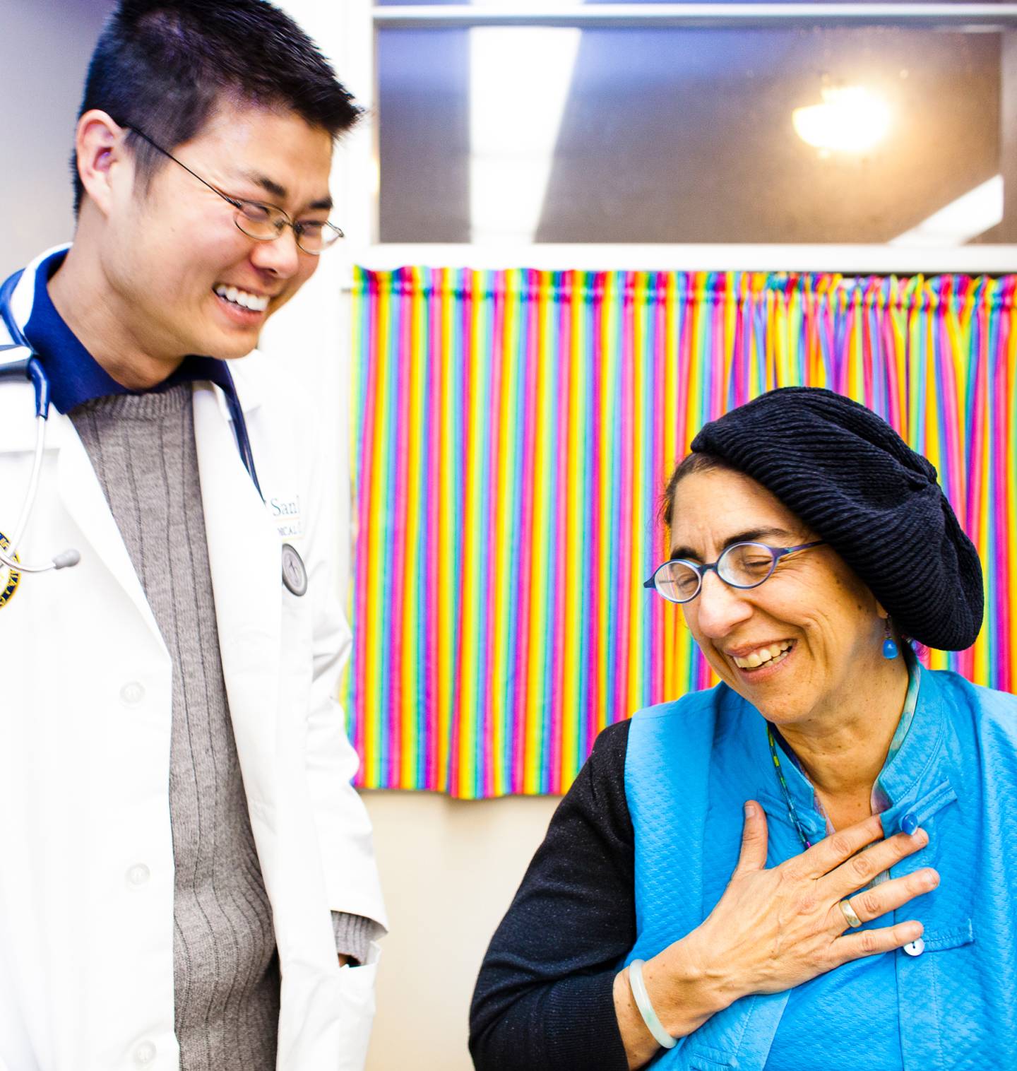 A doctor in a white coat with a stethoscope talks with a seated woman