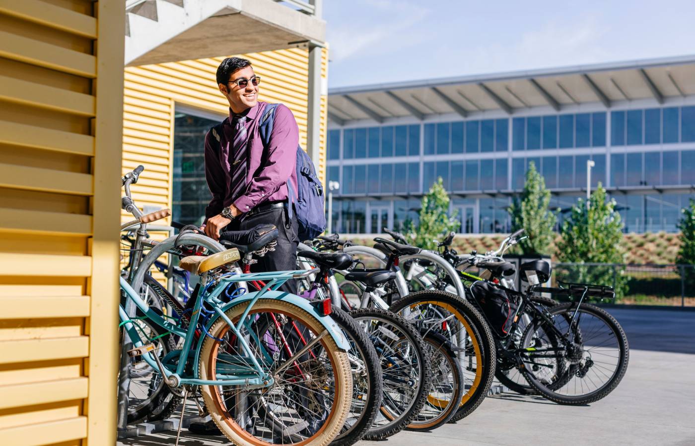 A young man in a purple shirt and sunglasses stands next to parked bicycles, smiling with a backpack over one shoulder.