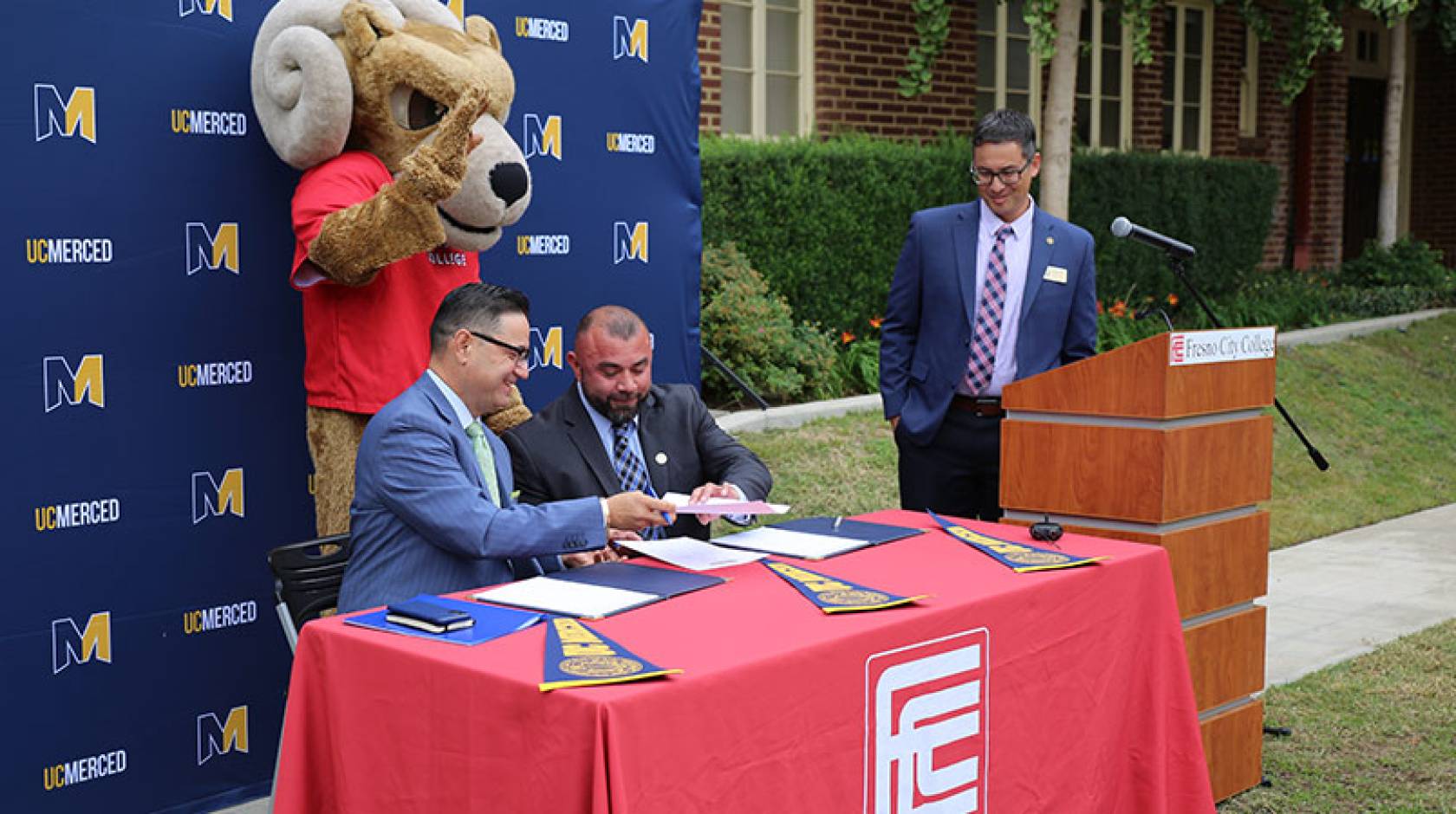 A ram mascot behind two men at a table passing a paper while a man at a lectern looks on