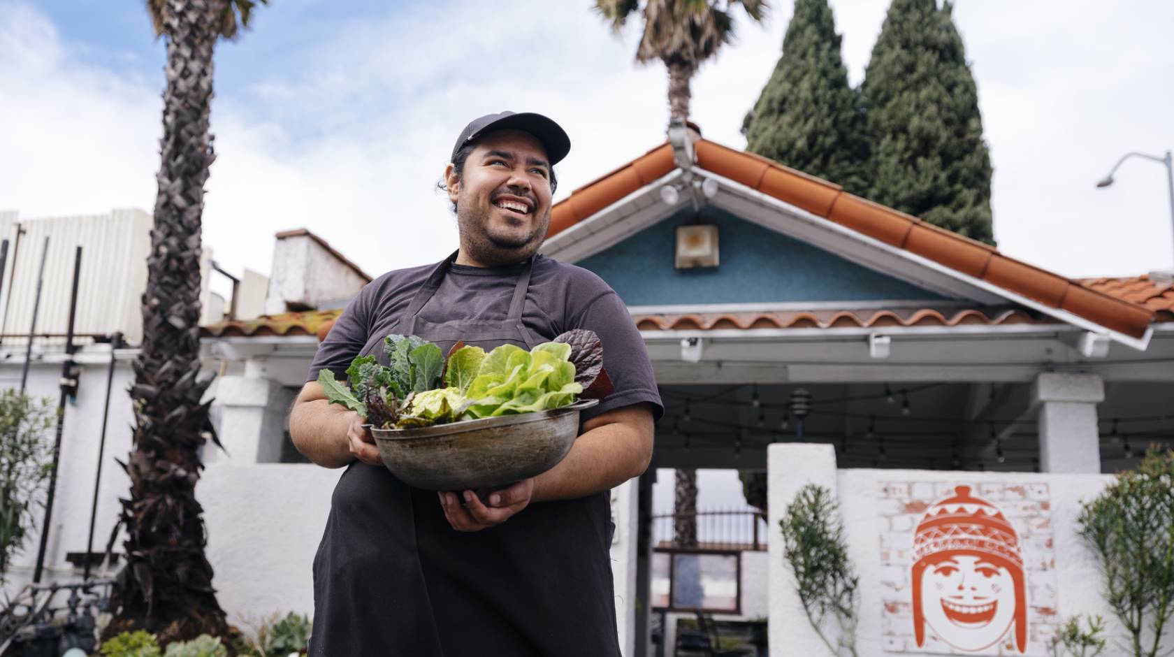 A young man smiles looking up and away from the camera, holding an armful of recently harvested lettuces. He's standing in front of a low green building with palm trees in the background. It looks like LA. 