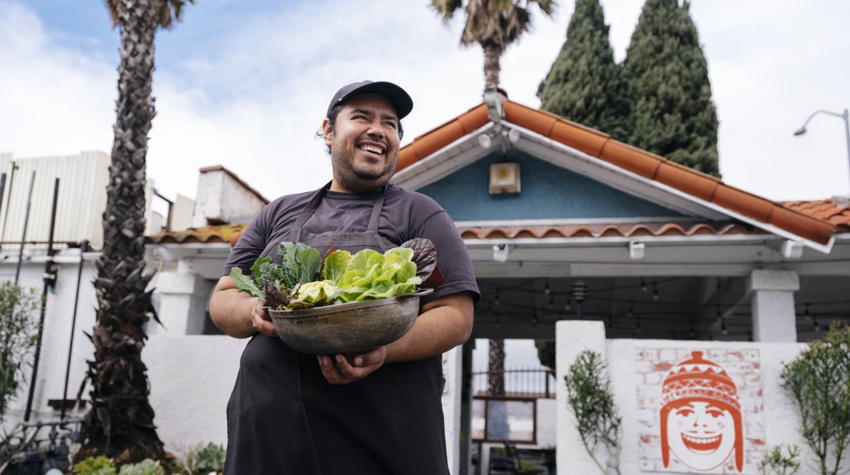 A young man smiles looking up and away from the camera, holding an armful of recently harvested lettuces. He's standing in front of a low green building with palm trees in the background. It looks like LA.