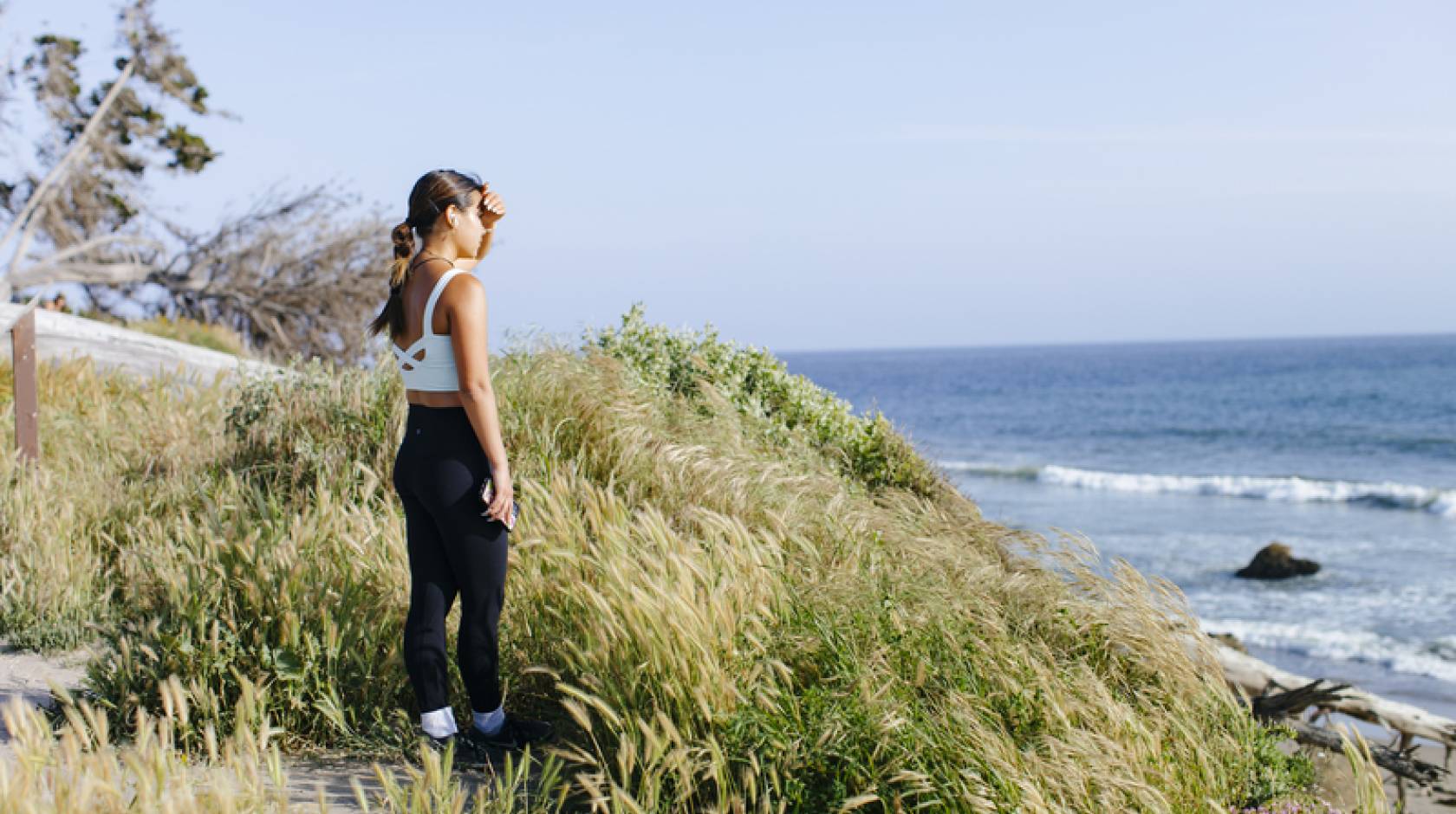 A young woman in exercise attire stands on a grassy bluff overlooking a beach, shading her eyes from the sun
