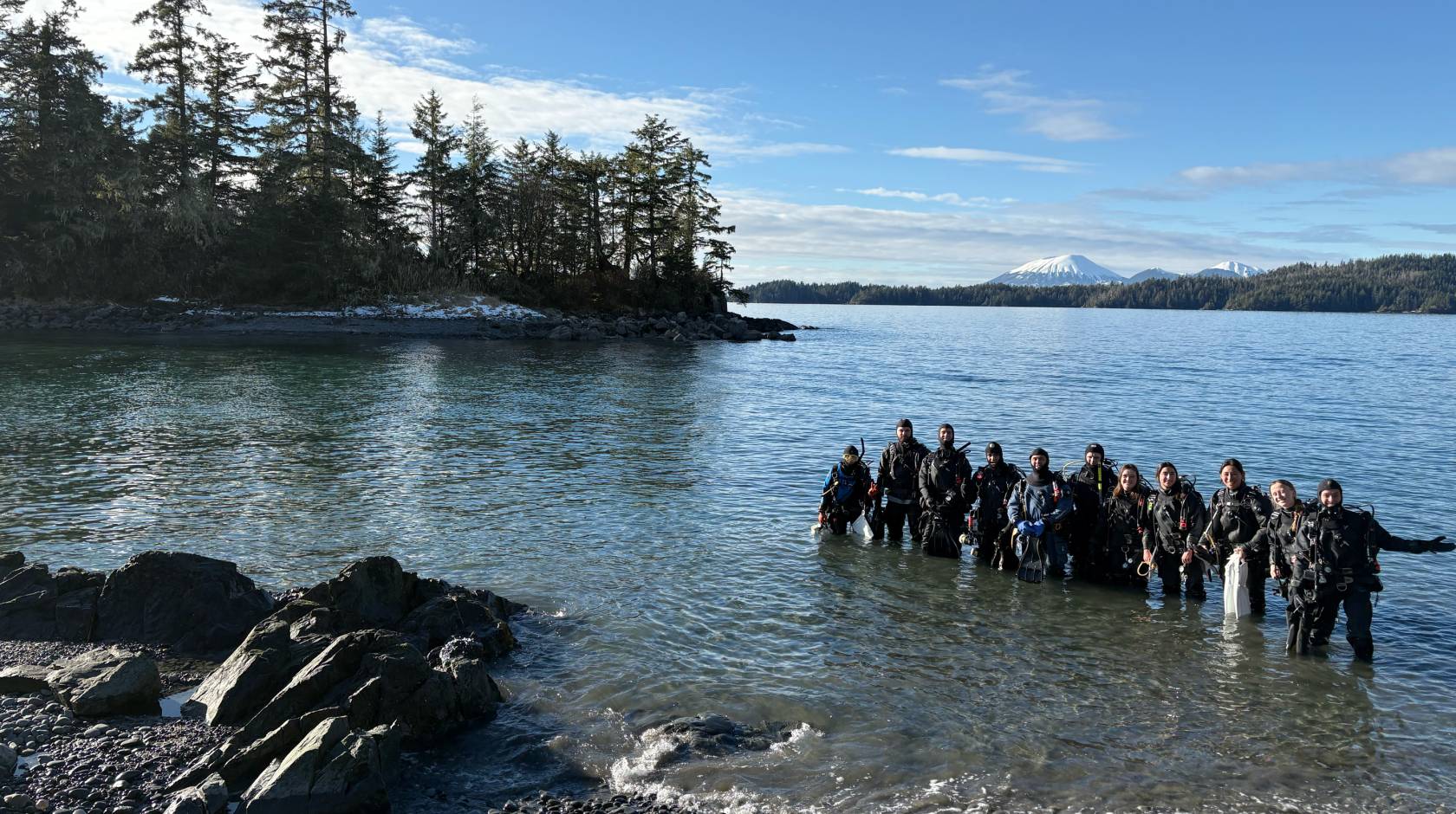 A smiling group of people stands in the water in wetsuits and SCUBA gear, with evergreen trees and a snow-covered peak in the background