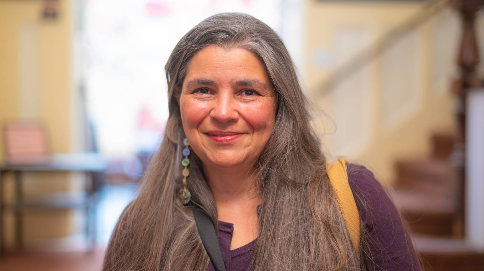 Carolyn Smith smiles at the camera in a shoulders-up portrait with a staircase and window in the background. She has long, straight, light-brown hair tinted with gray and wears a maroon top and long, dangly earrings. 