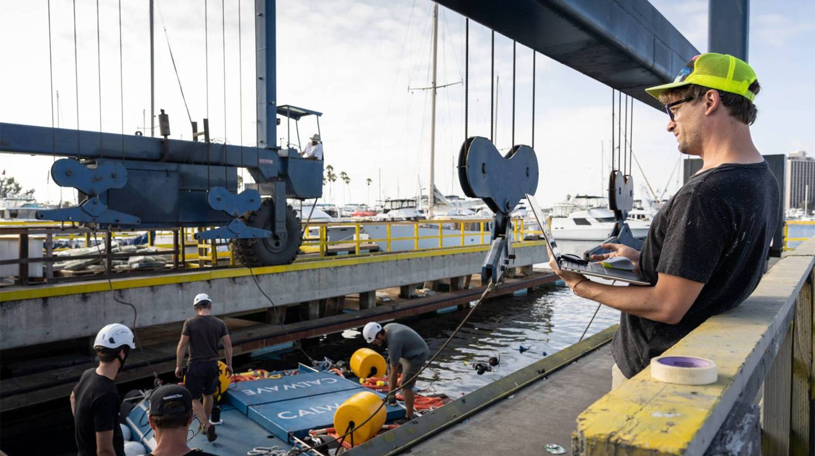 People in hard hats work under a crane on a dock, with a blue, car-sized platform in the middle of the frame