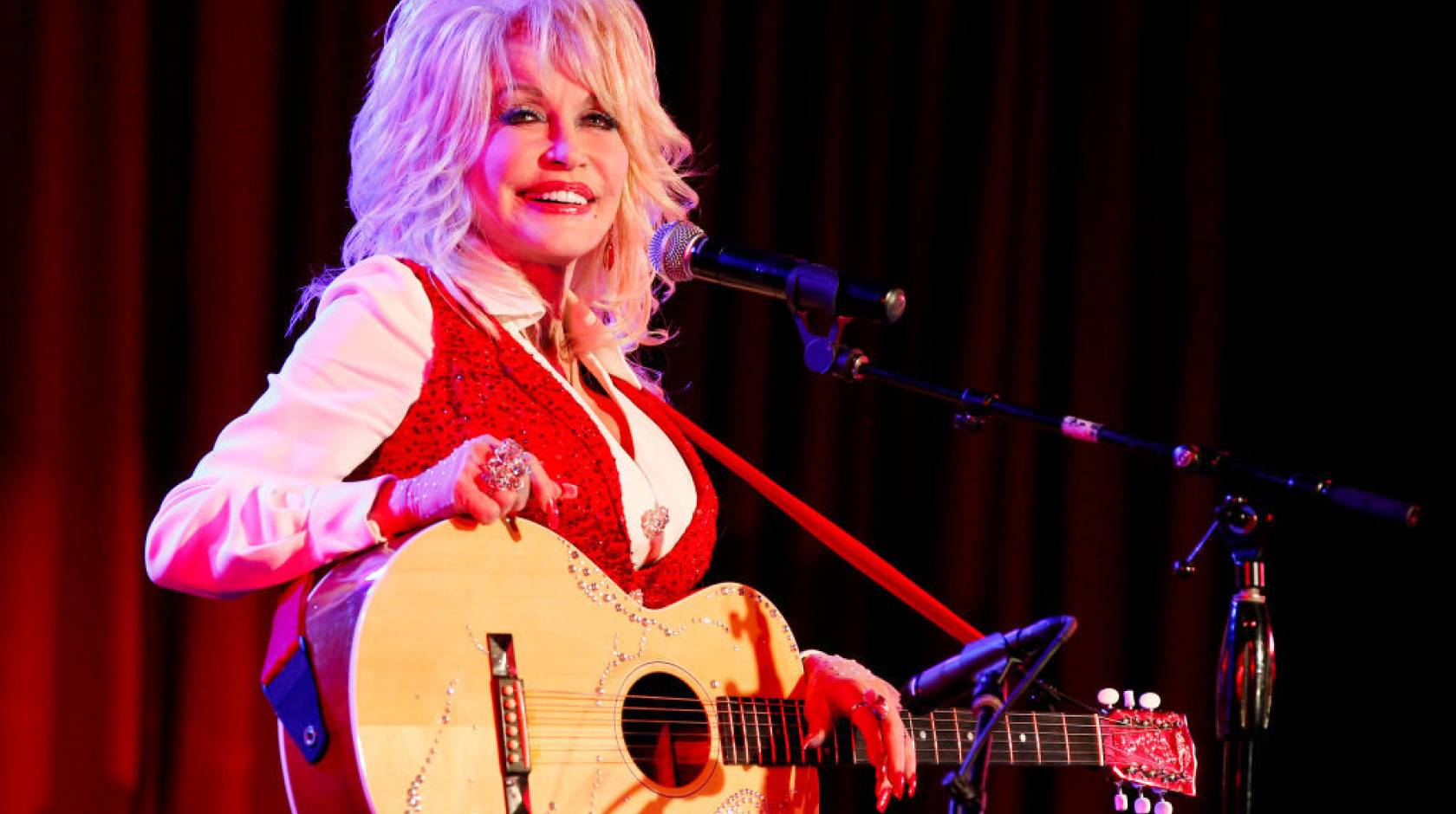 Dolly Parton in a red vest smiles while holding an acoustic guitar