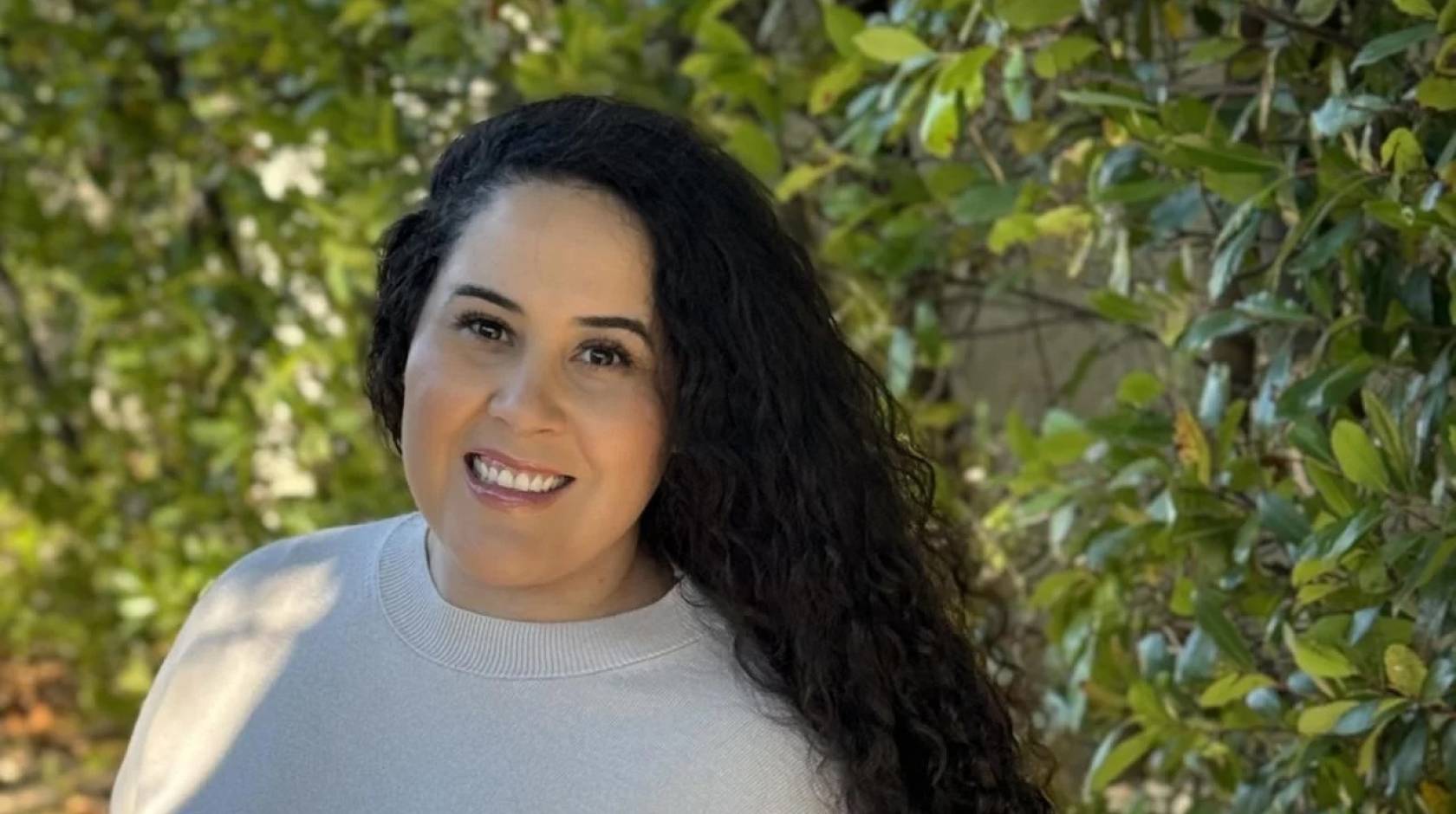 Lizbeth Lopez, 33, long curly dark hair, smiles in a shoulders-up portrait, with leaves in the background