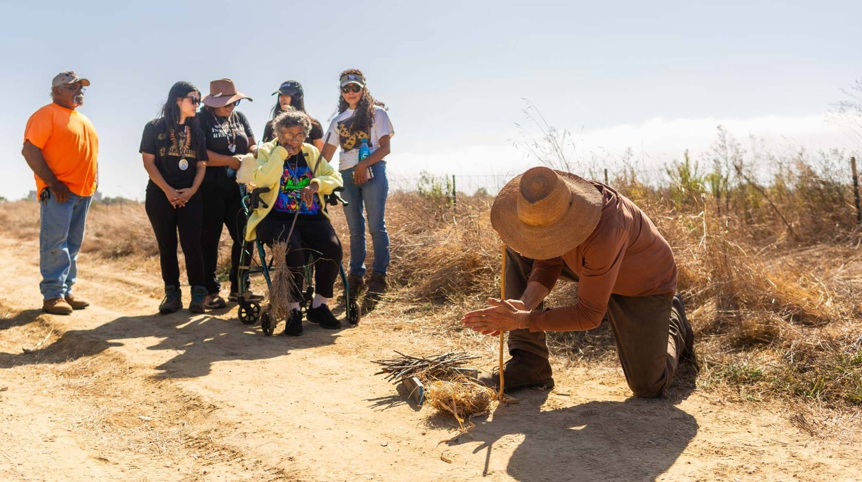 A man in a straw hat uses a wooden stick to start a fire in a brush pile while 6 people watch.