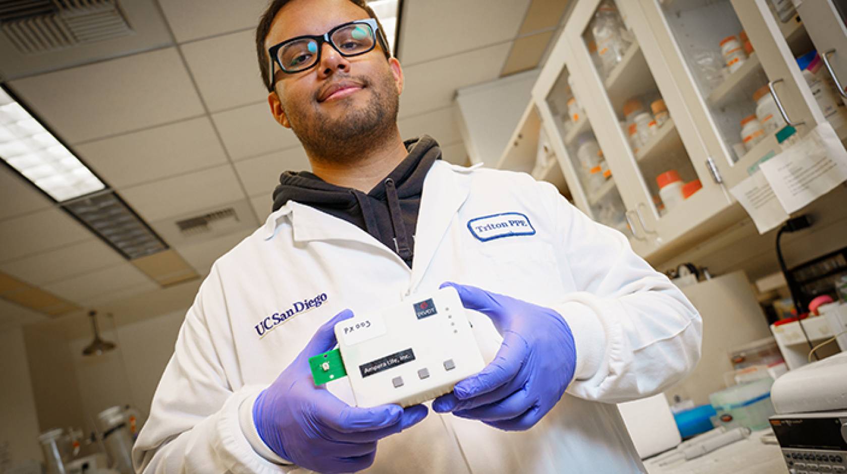 A male student in a white lab coat wearing glasses and purple rubber gloves holds a white box the size of a large sandwich in both hands