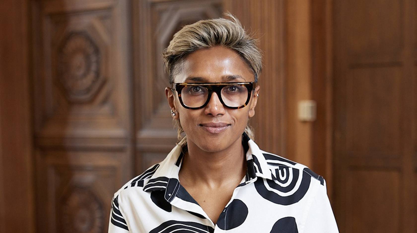 Poulumi Saha, wearing black rimmed glasses and a black and white blouse, smiles faintly for a shoulders-up portrait, with wood paneled walls in the background.