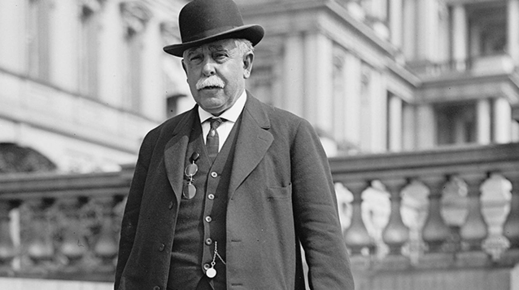 A man with gray hair in a bowler had and three-piece suit in front of a Neoclassical building